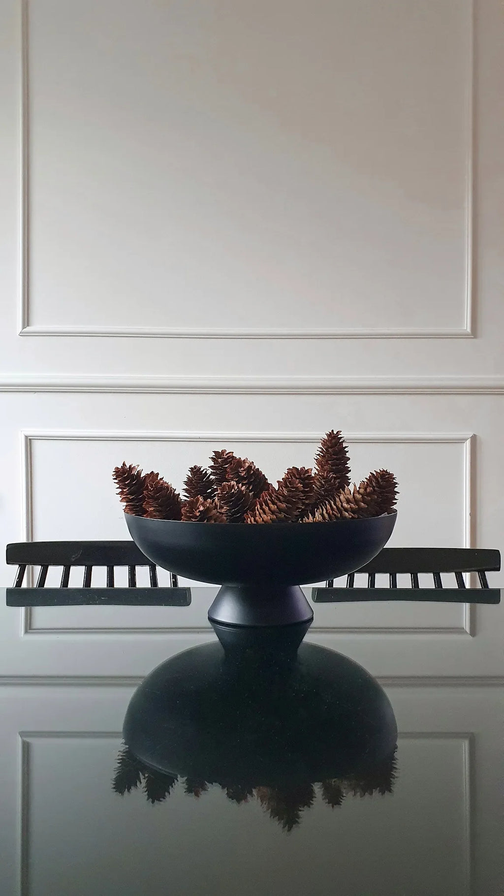 Decorative bowl with pine cones on a reflective surface against a white paneled wall.