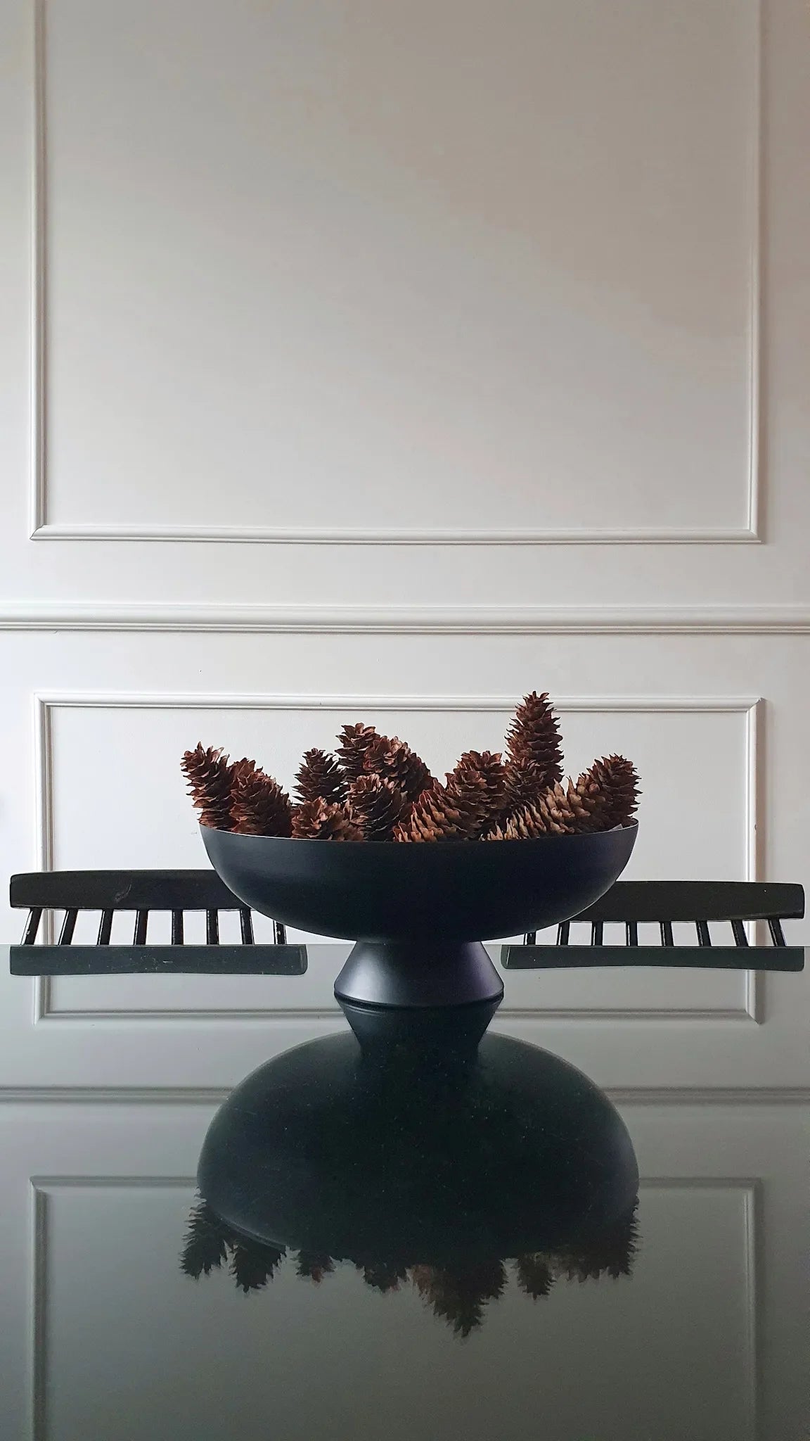 Decorative bowl with pine cones on a reflective surface against a white paneled wall.