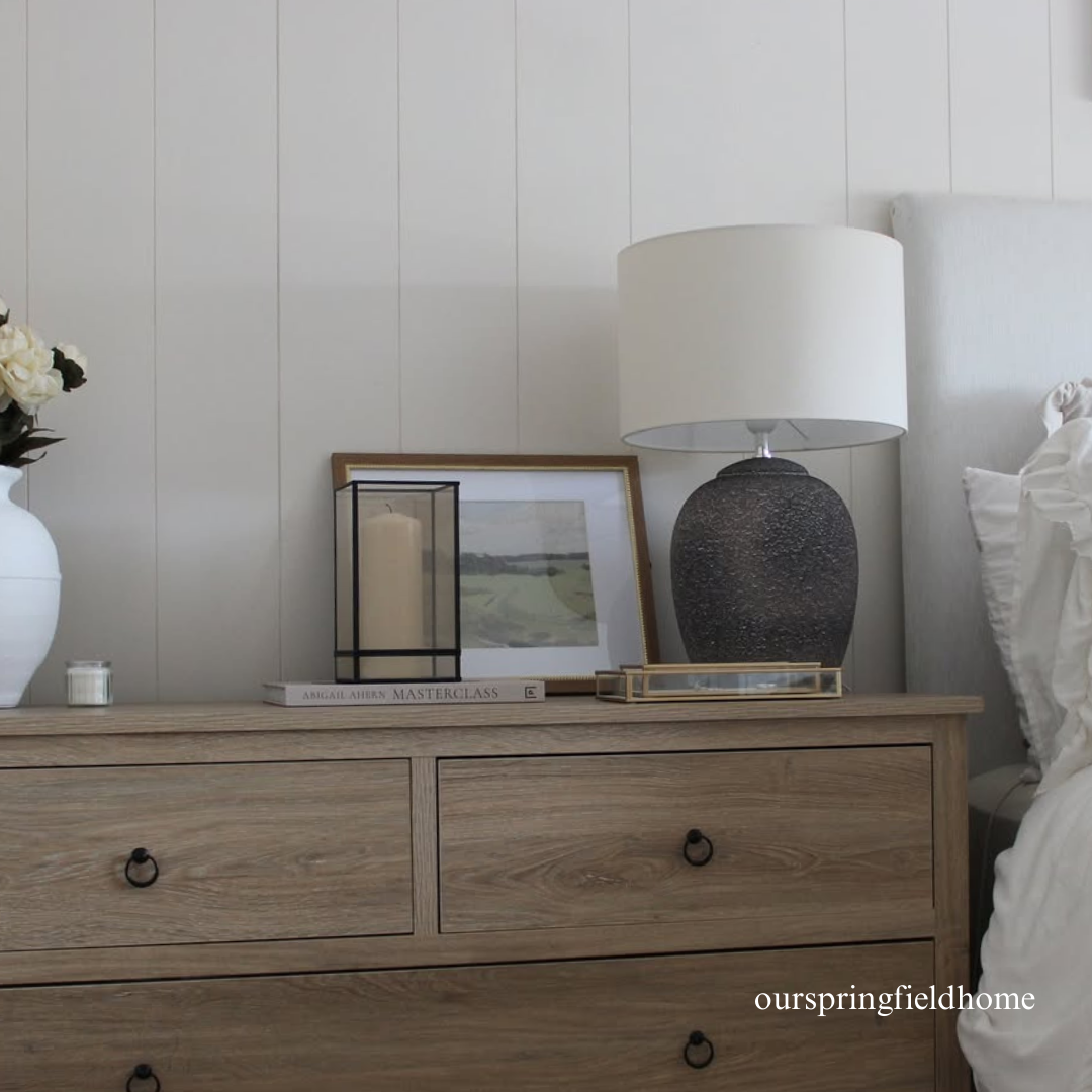 Wooden dresser with decorative items including a lamp, candles, and framed pictures against a white panelled wall.